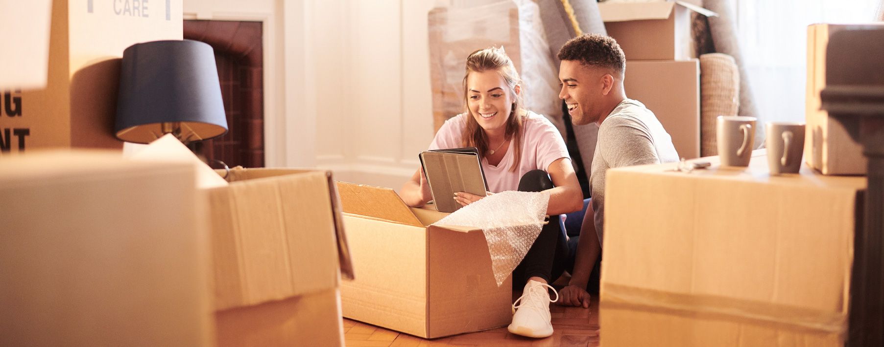 A young couple sits on the floor looking at a tablet surrounded by moving boxes