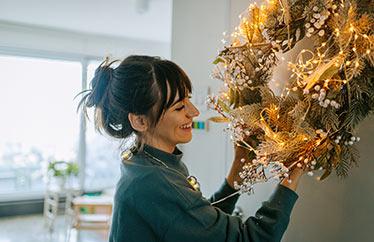 smiling homeowner putting up a lit holiday wreath in their home